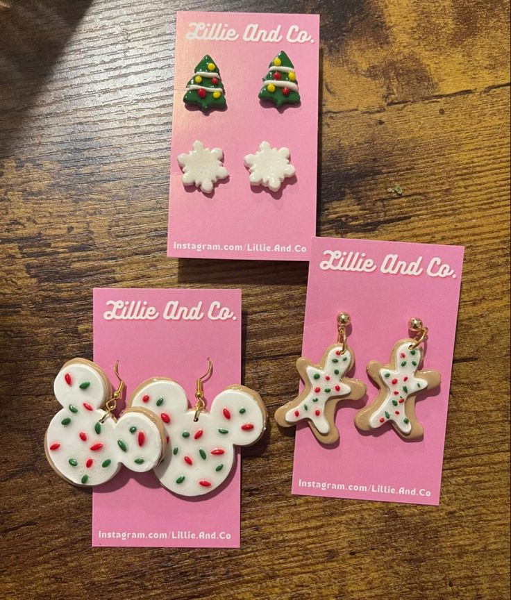 three christmas themed earrings on top of a wooden table next to pink cards with white and green designs