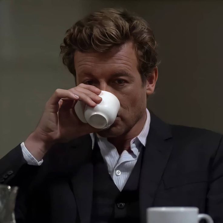 a man in a suit drinking out of a white coffee cup while sitting at a table