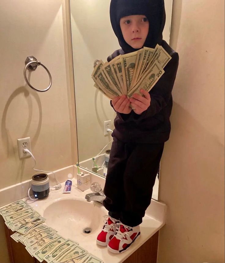 a little boy standing on top of a bathroom sink holding money in front of him