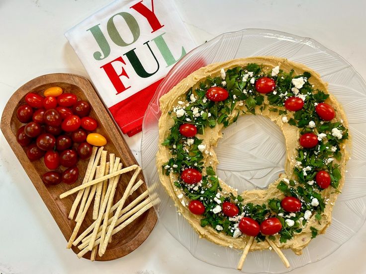 a christmas wreath made out of cheese, tomatoes and other toppings on a platter next to a box of joyful