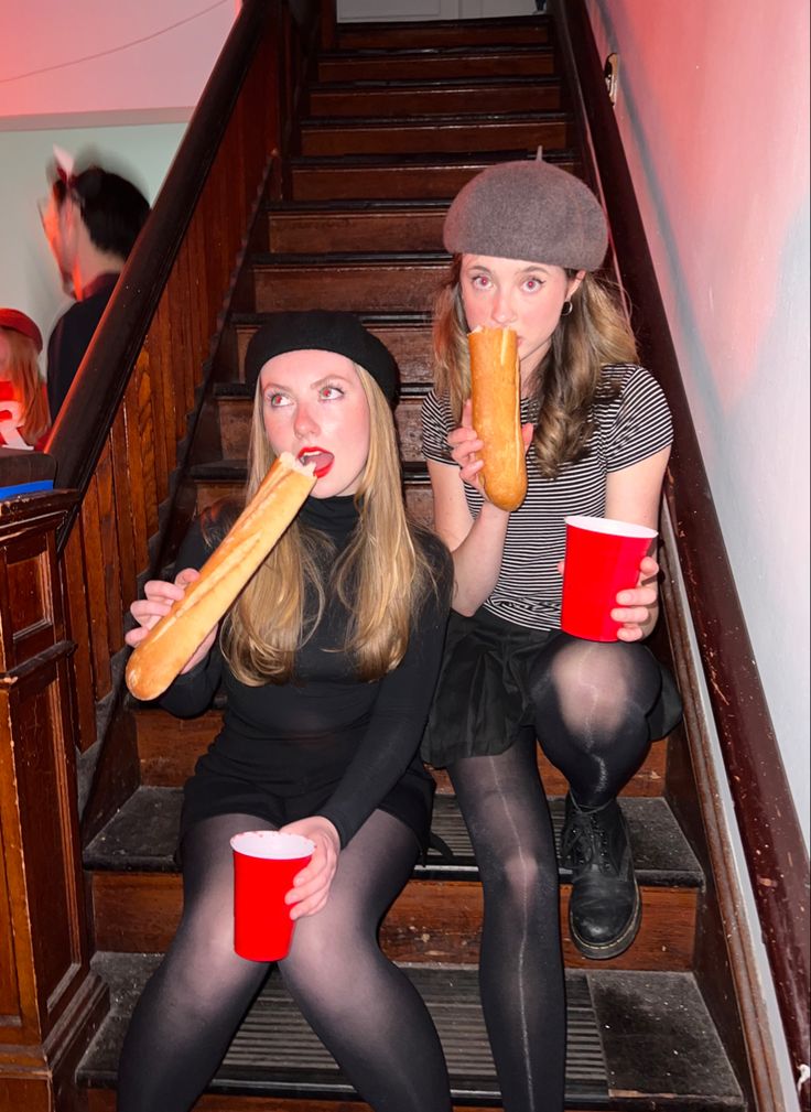 two women sitting on the stairs eating hot dogs and drinking sodas while holding red cups