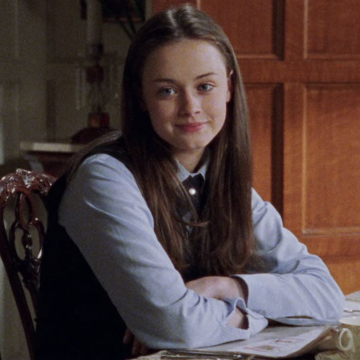 a woman sitting at a table with a book and pen in her hand while looking into the camera