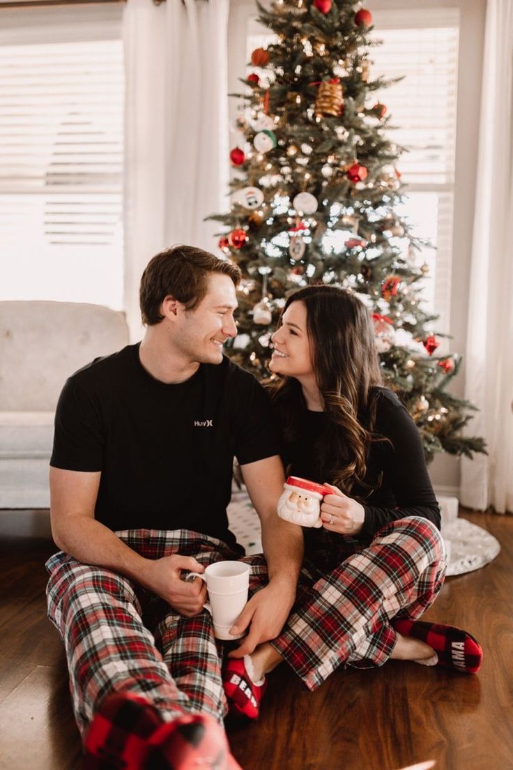 a man and woman sitting next to a christmas tree holding coffee cups in their hands