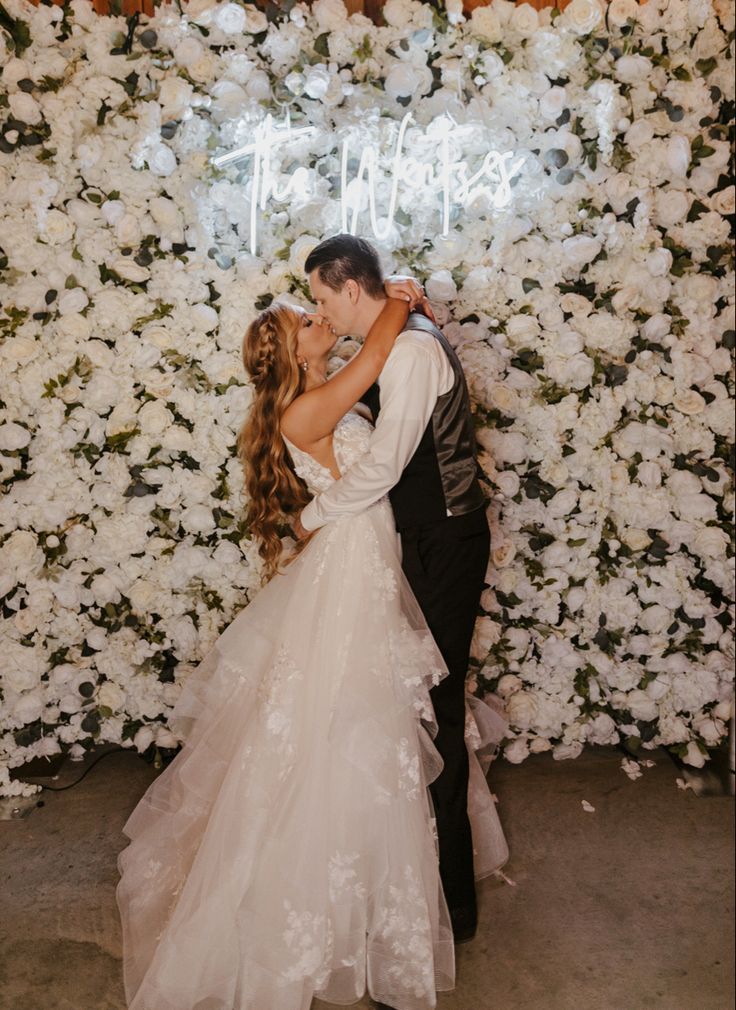 a bride and groom kissing in front of a flower wall with the words thank you written on it