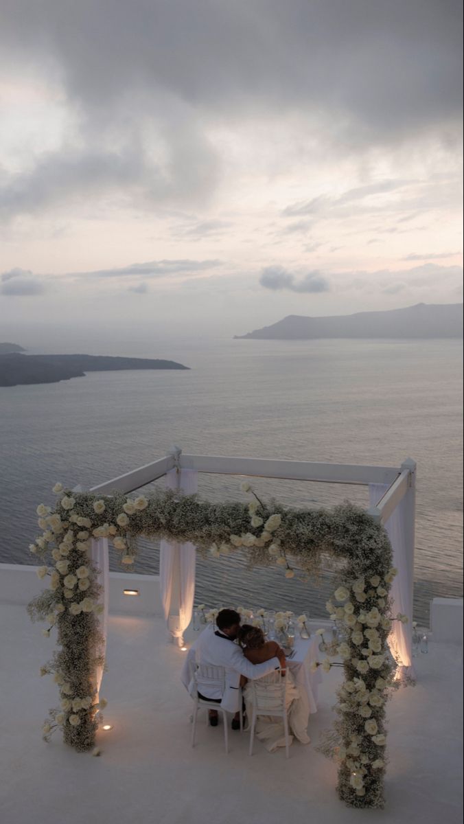 two people sitting at a table under an arch with flowers on it and the ocean in the background