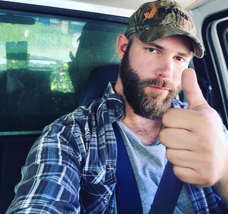 a man with a beard wearing a camo hat and holding his thumb up to the camera