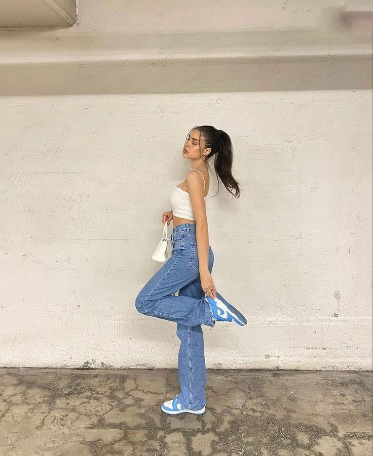 a woman standing in front of a white wall with her hand on her hip and wearing blue jeans