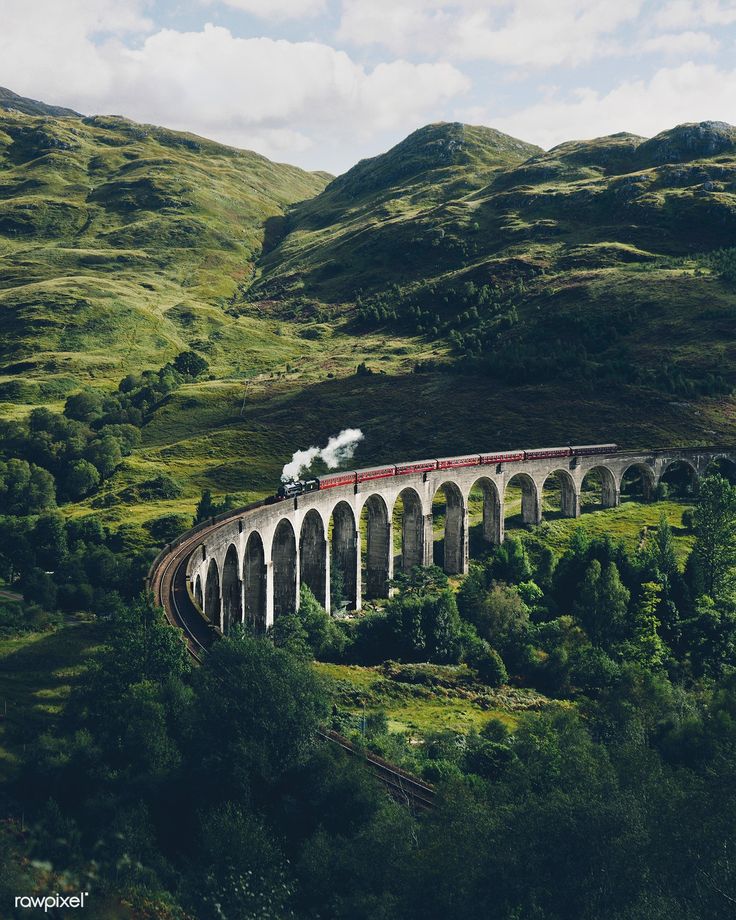 a train traveling over a bridge on top of a lush green hillside next to mountains