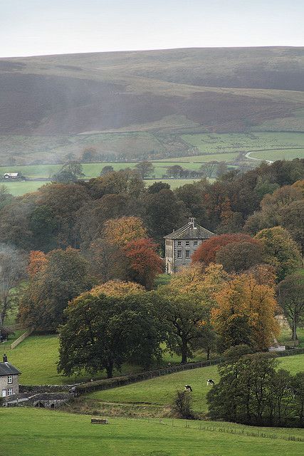 an old house surrounded by trees in the middle of a green field with hills in the background