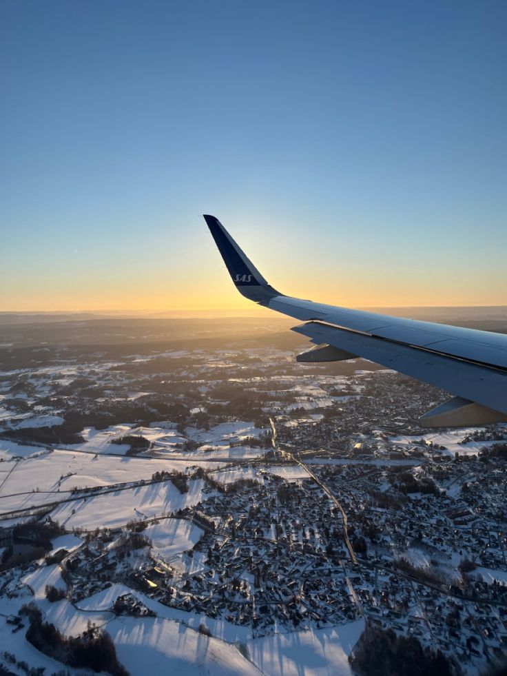 the wing of an airplane as it flies over snow - covered fields and trees at sunset