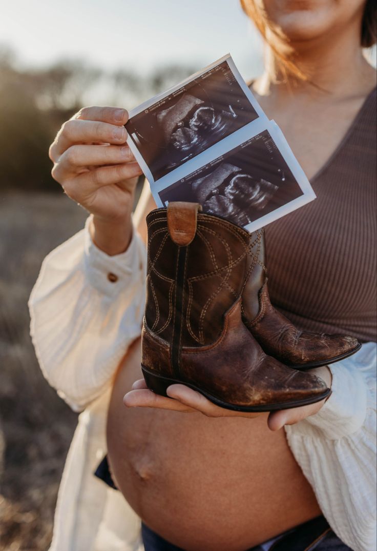 a pregnant woman holding up an old pair of cowboy boots to show off her baby bump