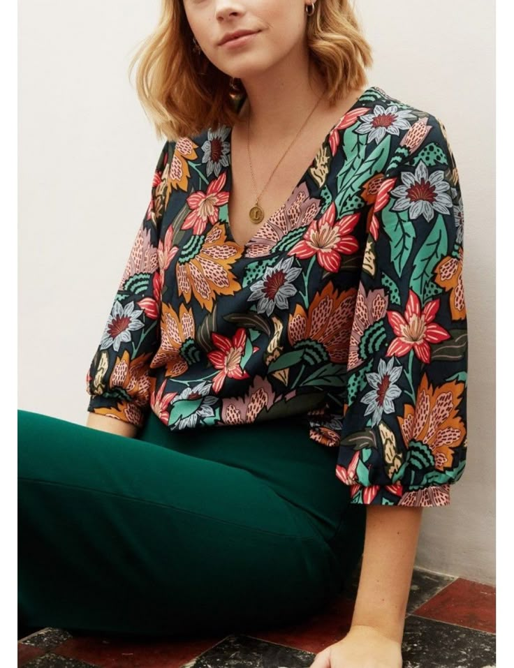 a woman sitting on the ground wearing green pants and a floral shirt with her hands in her pockets