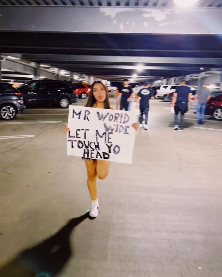 a woman holding a sign that says mr world wide let me touch to head in an empty parking lot