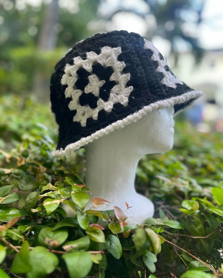 a white mannequin head wearing a black and white crocheted hat on top of green bushes