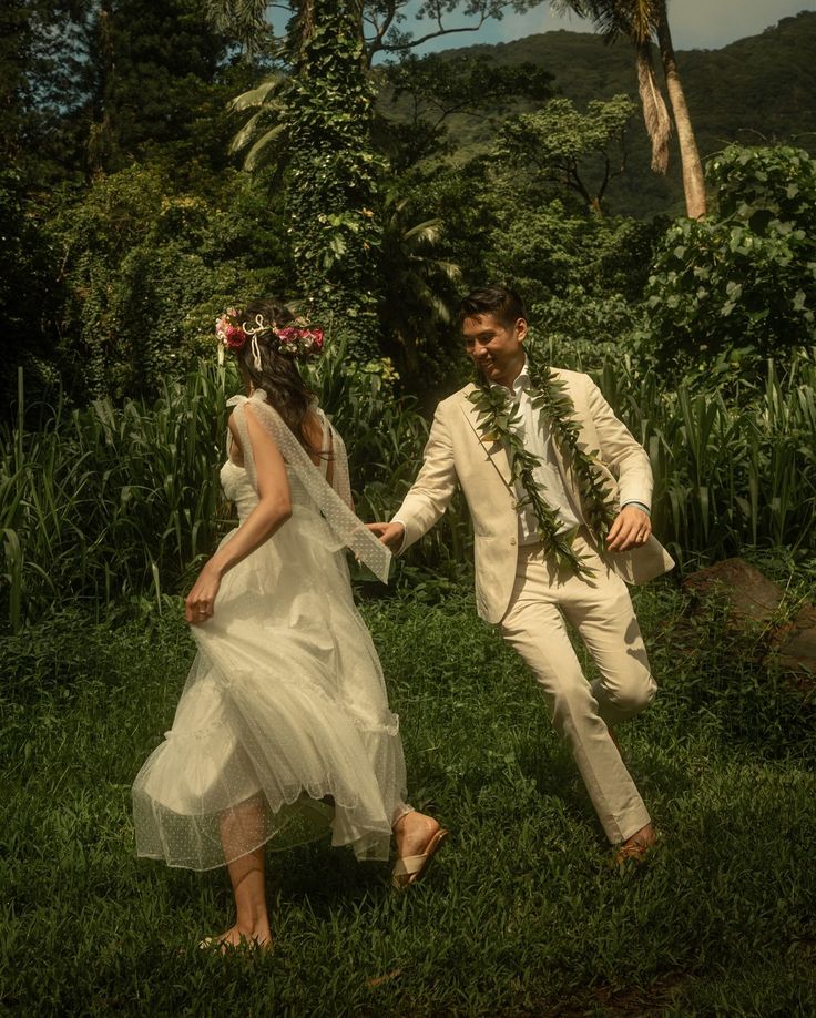 a man and woman dressed in white walking through the grass with flowers on their head