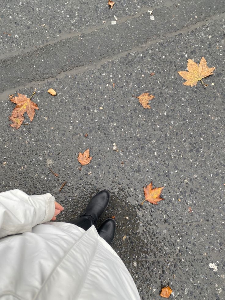 a person in white coat and black shoes standing on sidewalk next to leafy ground