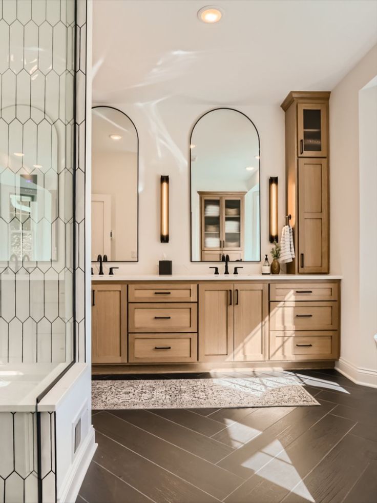 Go to the link to see more of this stunning primary bathroom remodel. This custom vanity was built for beauty and tons of storage. Long arched mirrors and linear sconces. Black matte plumbing fixtures. Open glass doors on the top cabinets to display decor. Modern Scandinavian style. Black wood herringbone tile floors Vanity With Black Tile Floor, Master Bath Long Vanity, Custom Double Vanity Bathroom, Primary Bathroom Double Vanity, Arched Mirror With Sconces, 8ft Bathroom Vanity Master Bath, Bathrooms With Matte Black Fixtures, Tile For Master Bath Floor, Lightwood Bathroom Vanity