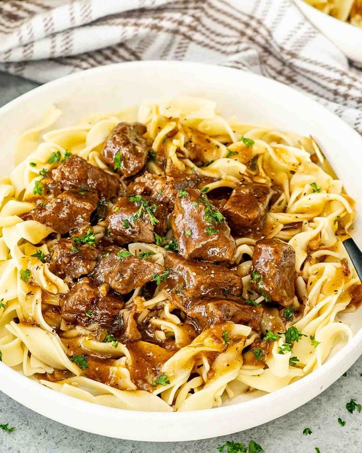 a white bowl filled with beef and noodles on top of a marble table next to a fork