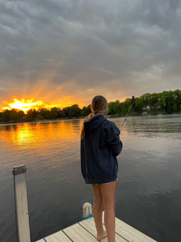 a woman standing on a dock fishing at sunset