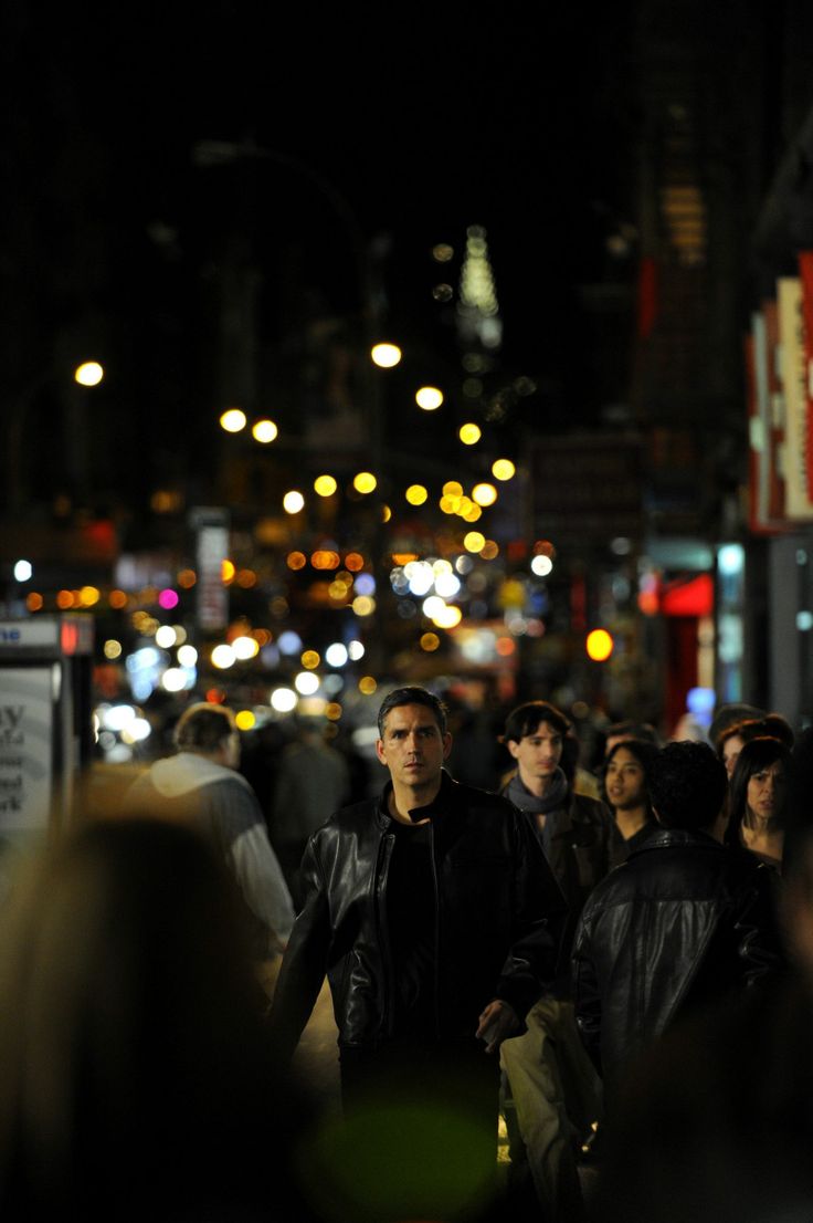 a group of people walking down a street at night time with lights in the background