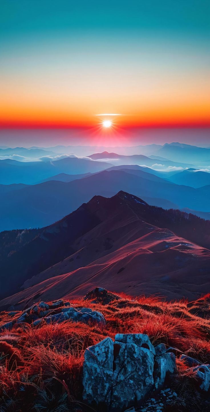 the sun is setting on top of a mountain with red grass and rocks in the foreground