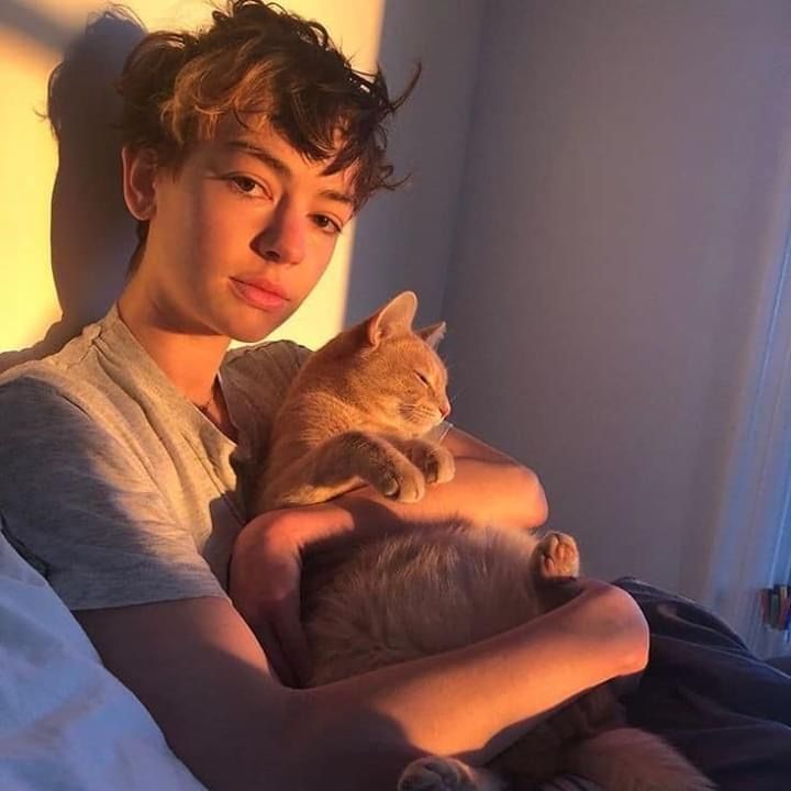 a young man holding a cat in his arms while sitting on a bed with sunlight coming through the window