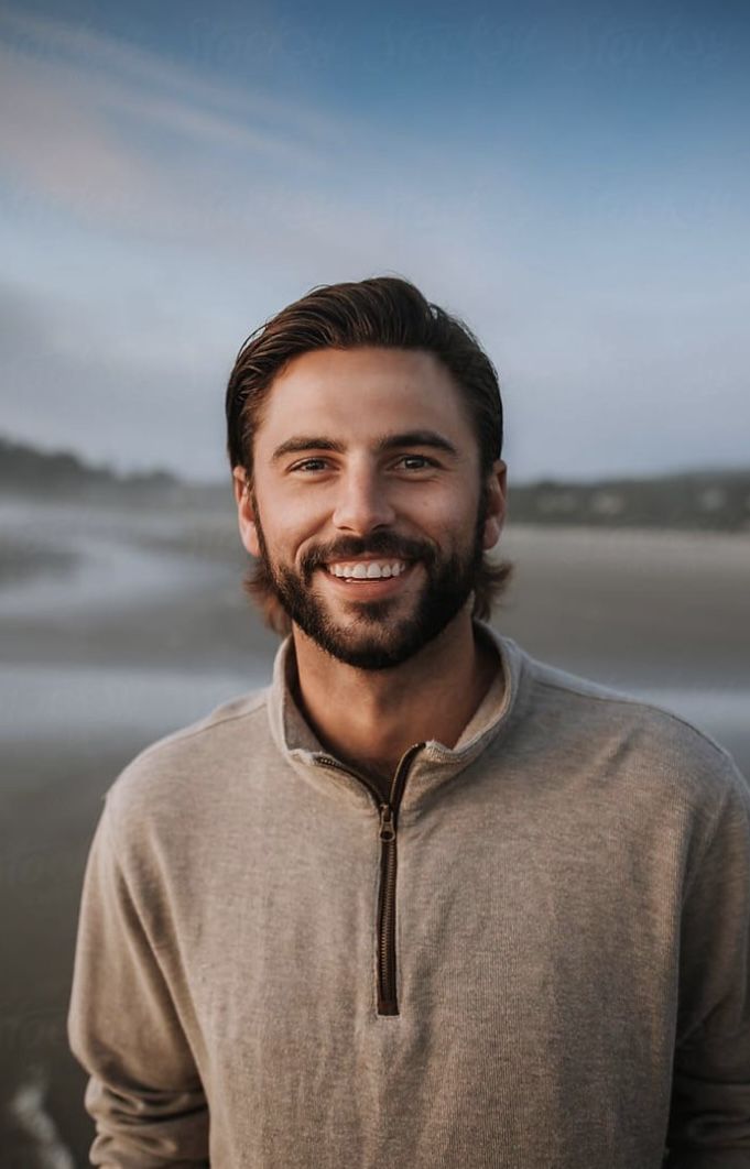 a man with a beard standing in front of the ocean smiling at the camera while wearing a sweater