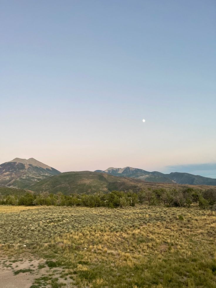 an empty field with mountains in the distance and grass on the ground, under a clear blue sky