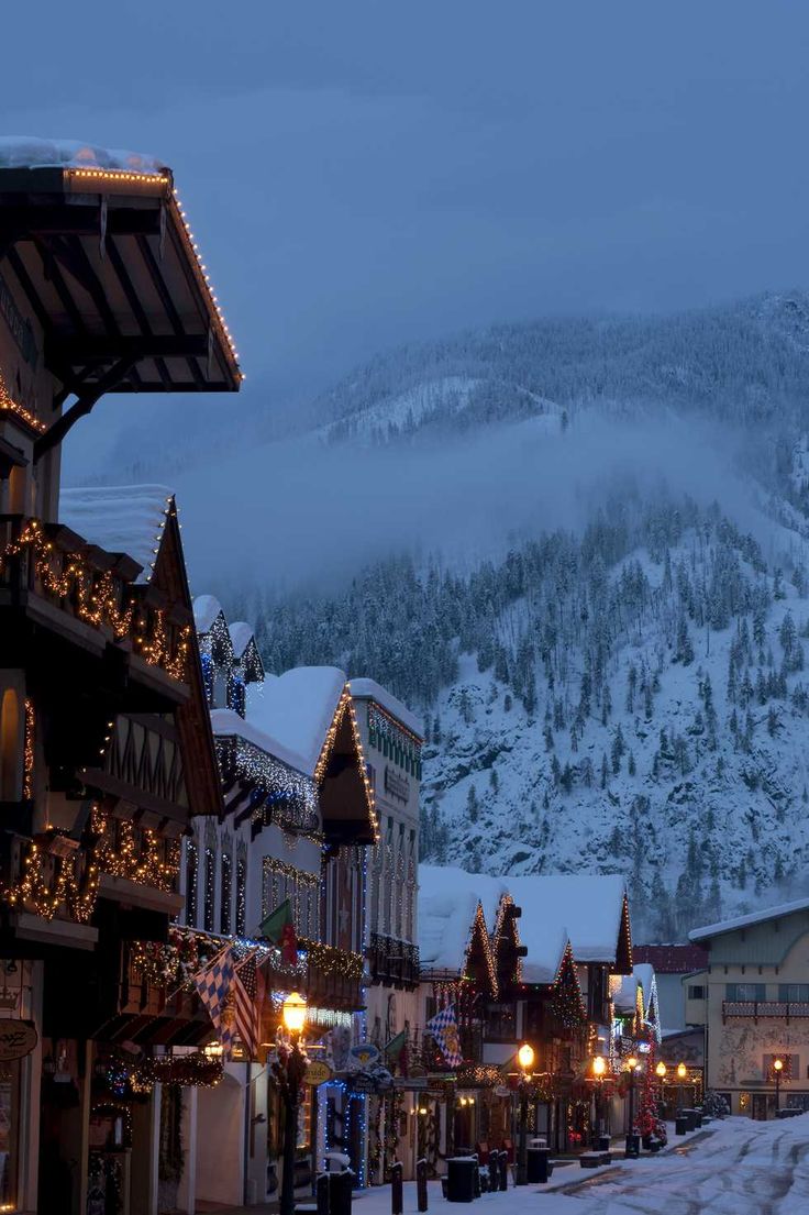 the town is decorated for christmas with lights and snow on the mountain behind it in the evening