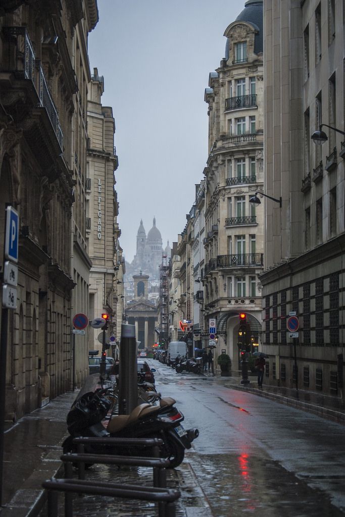 an empty city street in the rain with buildings on both sides and motorcycles parked along the sidewalk