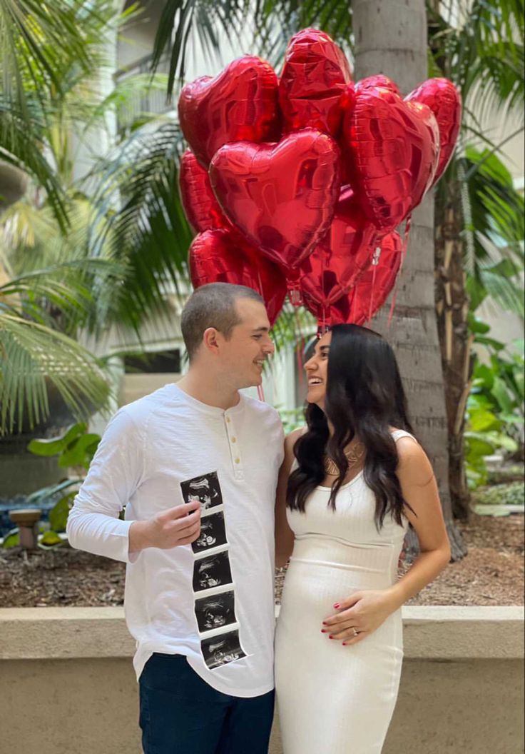 a man and woman standing next to each other holding red heart balloons