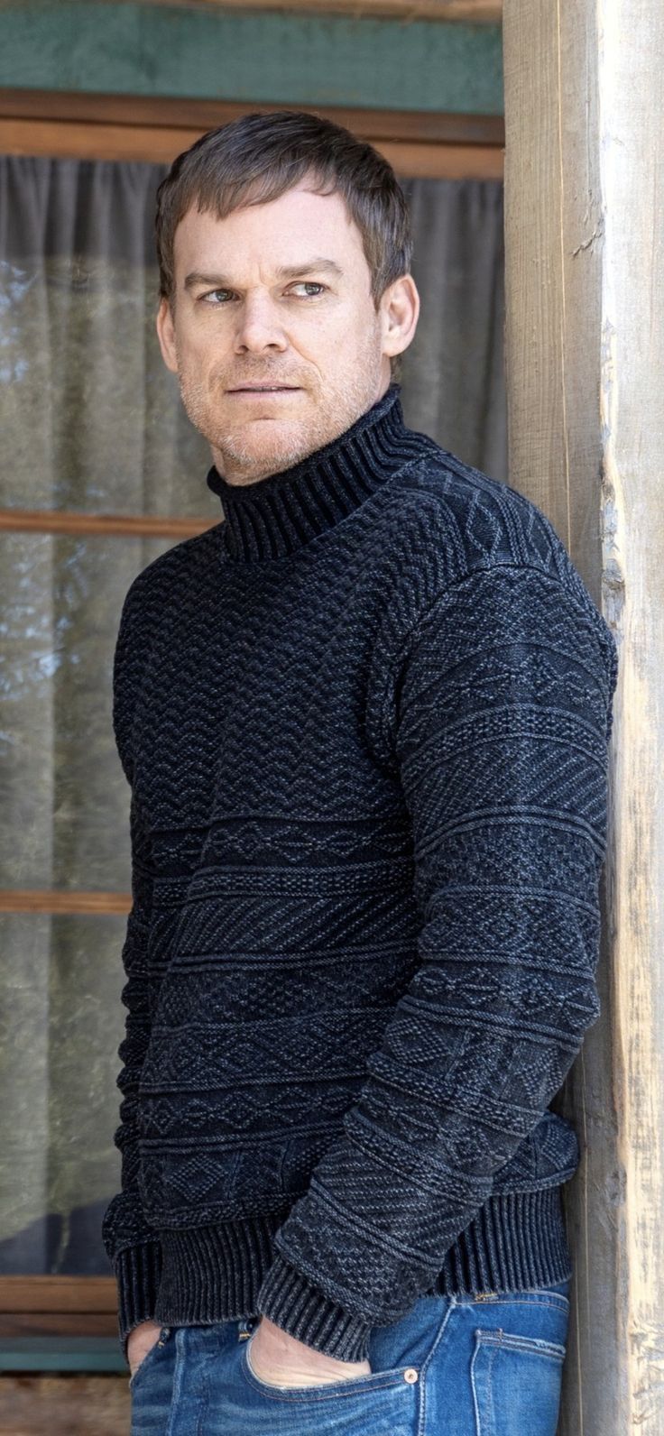 a man standing next to a wooden pole wearing a black turtle neck sweater and jeans