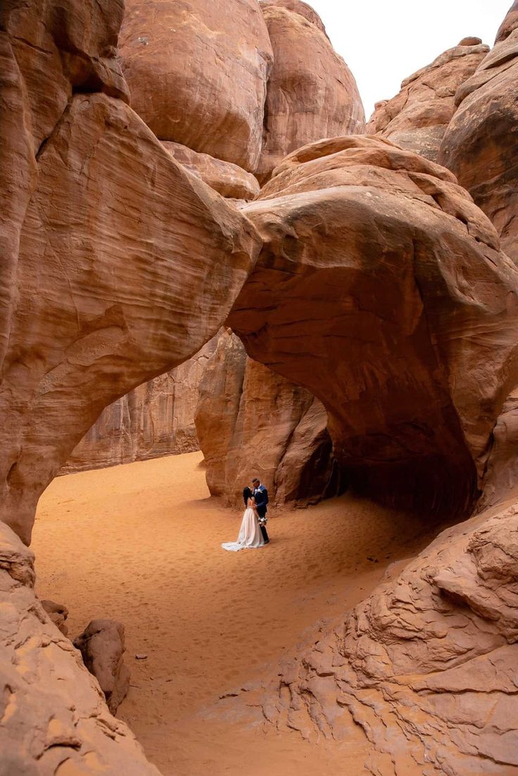 a man and woman standing in the middle of a desert area with large rock formations