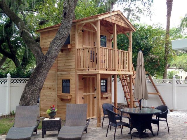 a small wooden house with a deck and patio furniture in the back yard, next to a large tree