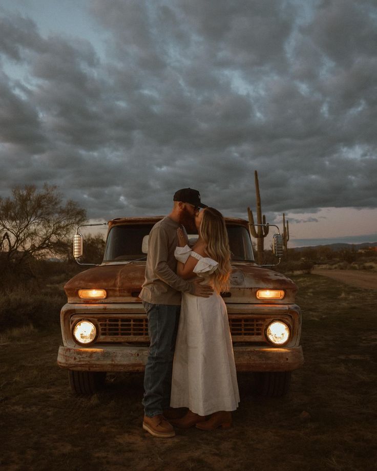 a man and woman kissing in front of an old pickup truck at dusk with clouds overhead