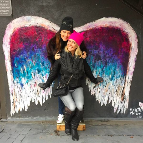 two women are posing in front of a wall with wings painted on it and one is hugging the other