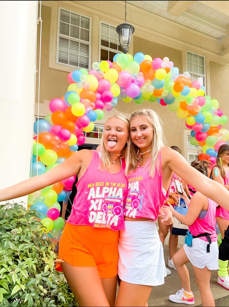 two girls in pink shirts and orange shorts are standing under a arch made out of balloons