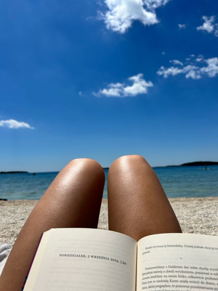 a person laying down reading a book on the beach with their feet up in the sand