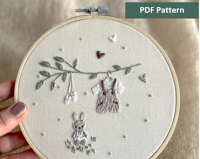 someone is holding up a hand embroidery pattern on a white background with the image of two rabbits hanging from a tree branch