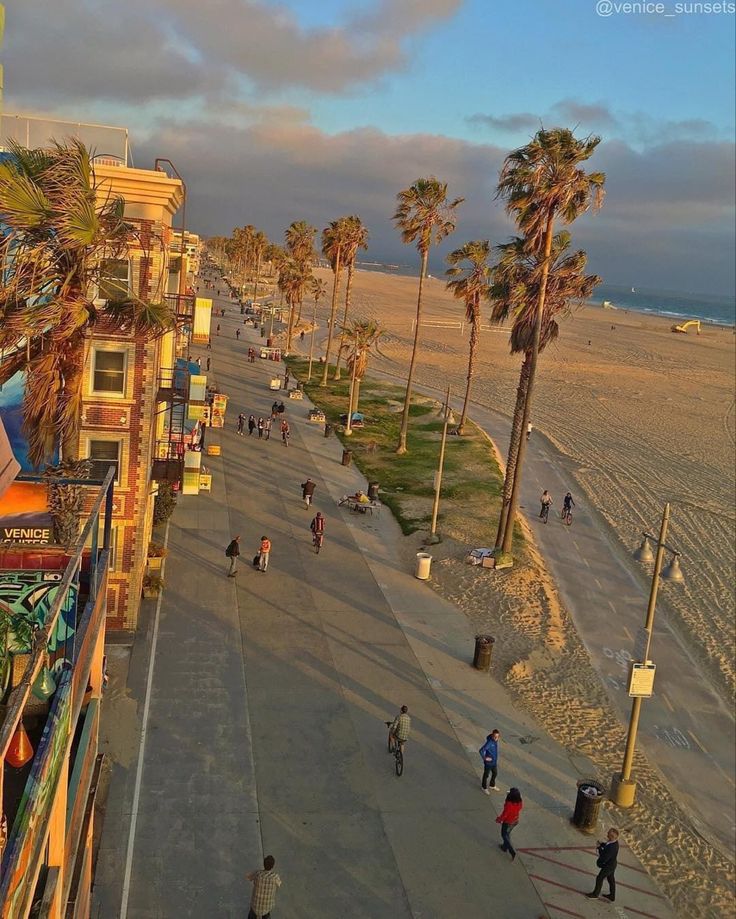 people walking and riding bikes on the beach near buildings with palm trees in the foreground