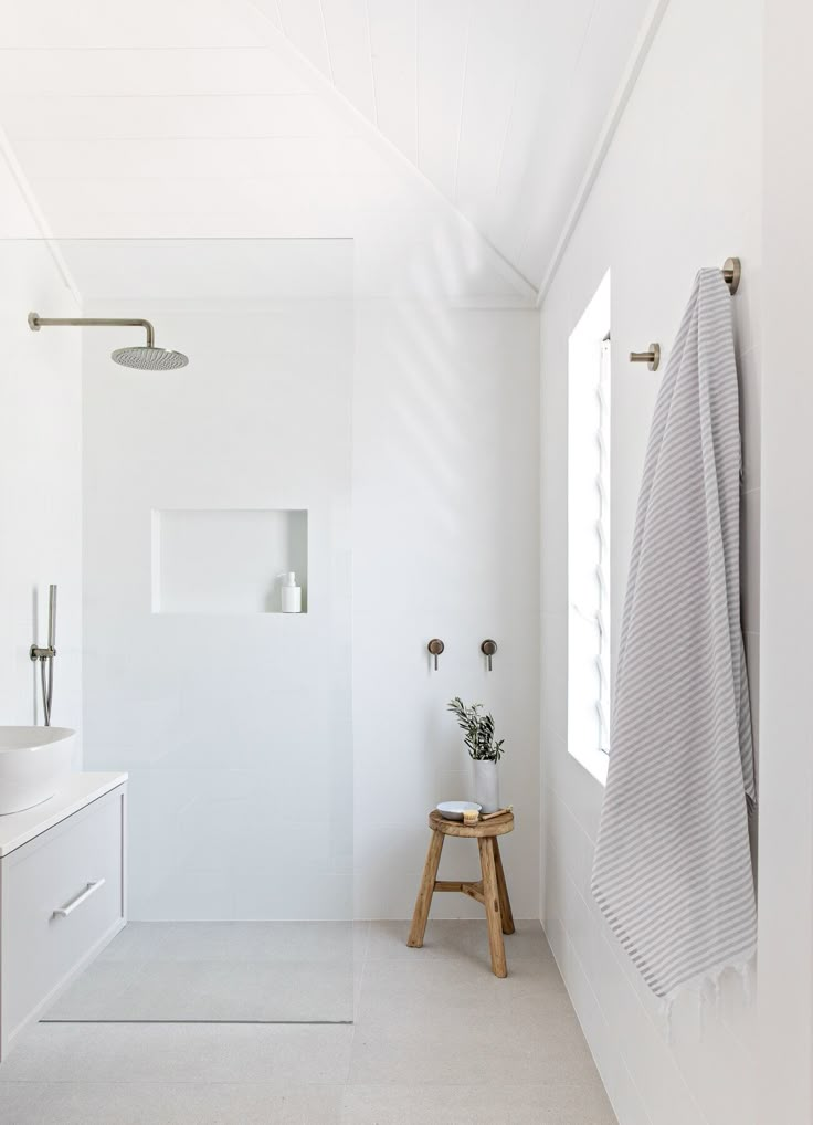 a bathroom with white walls and flooring, including a wooden stool next to the shower