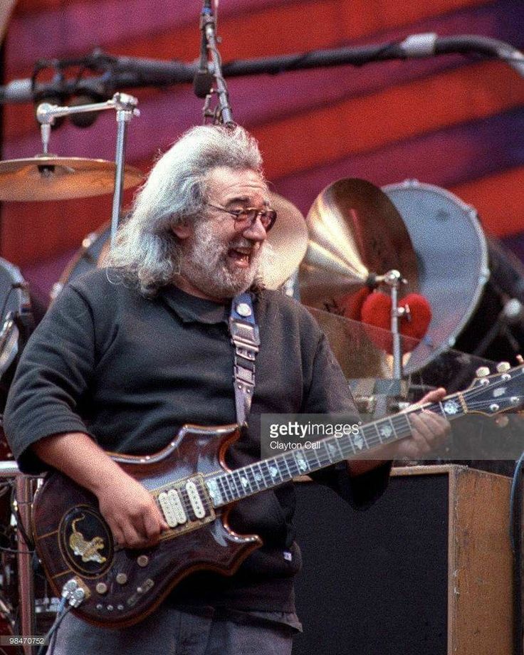 an older man with grey hair and glasses playing guitar in front of drums on stage