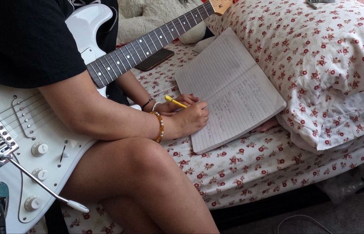 a person sitting on a bed with a guitar and notebook in front of their face