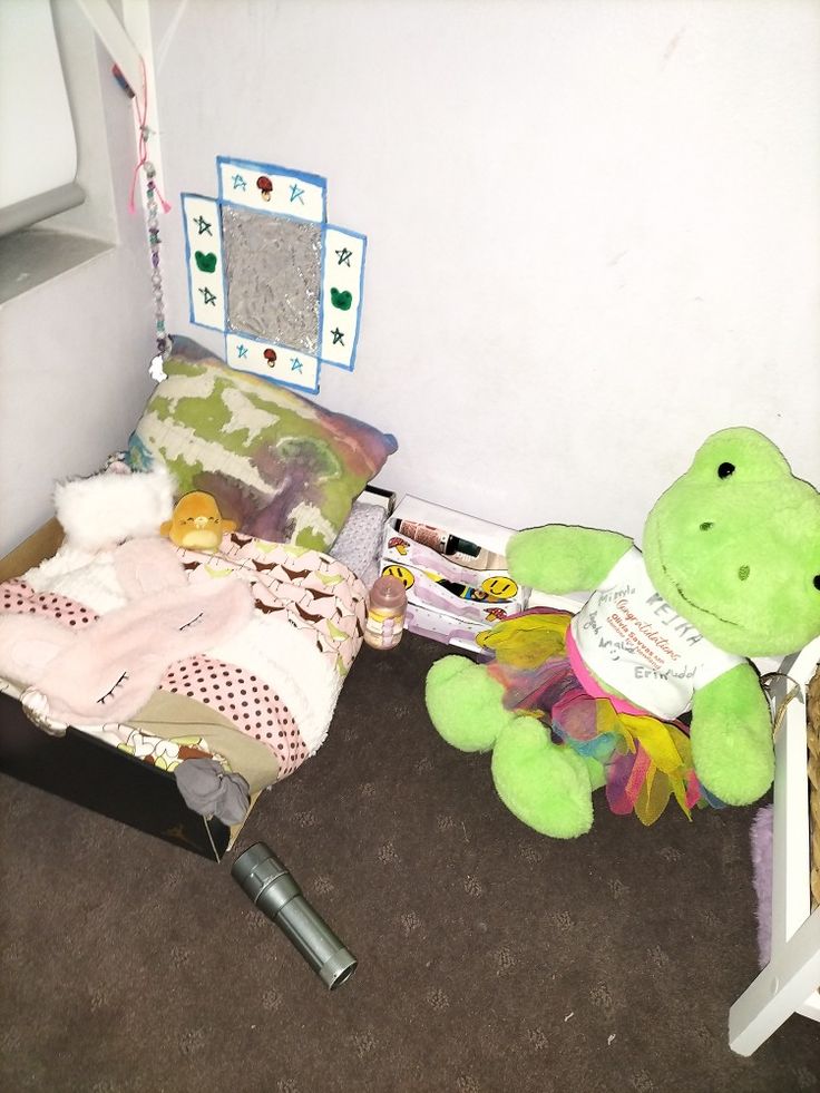 a green teddy bear sitting on the floor next to a crib with blankets and toys