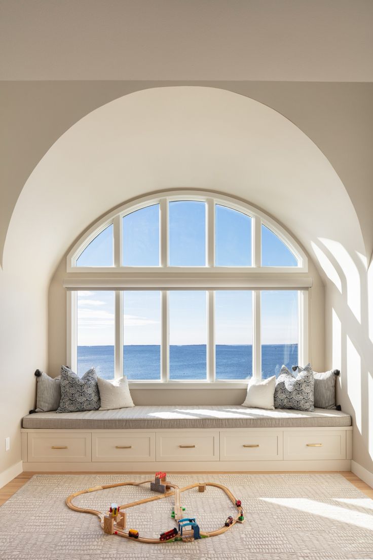 a window seat with toys on the floor in front of it and an arched window overlooking the ocean