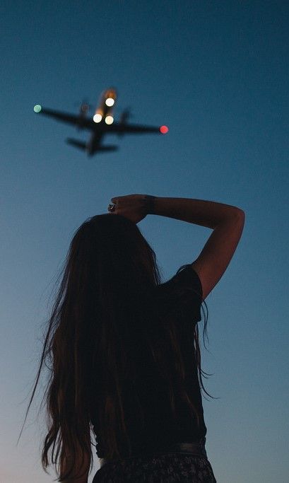 a woman looking at an airplane flying in the sky with her hair back to the camera