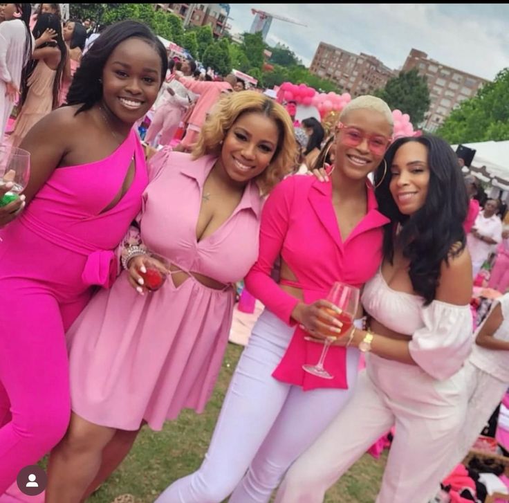 four women in pink dresses posing for a photo at an outdoor event with other people