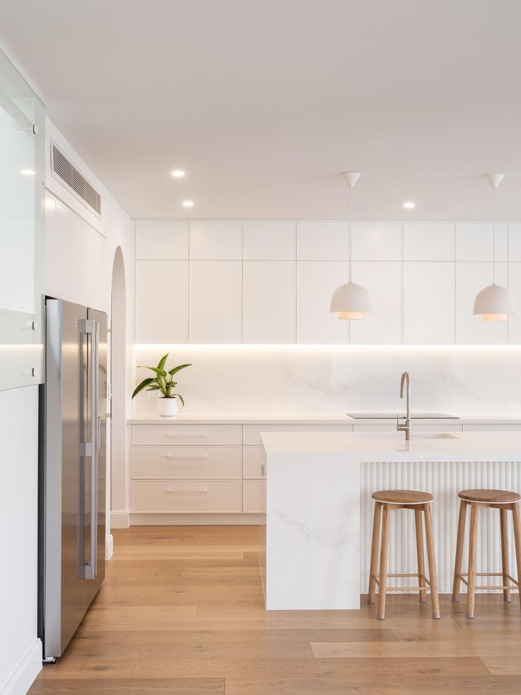 a white kitchen with two stools next to an island and refrigerator in the middle