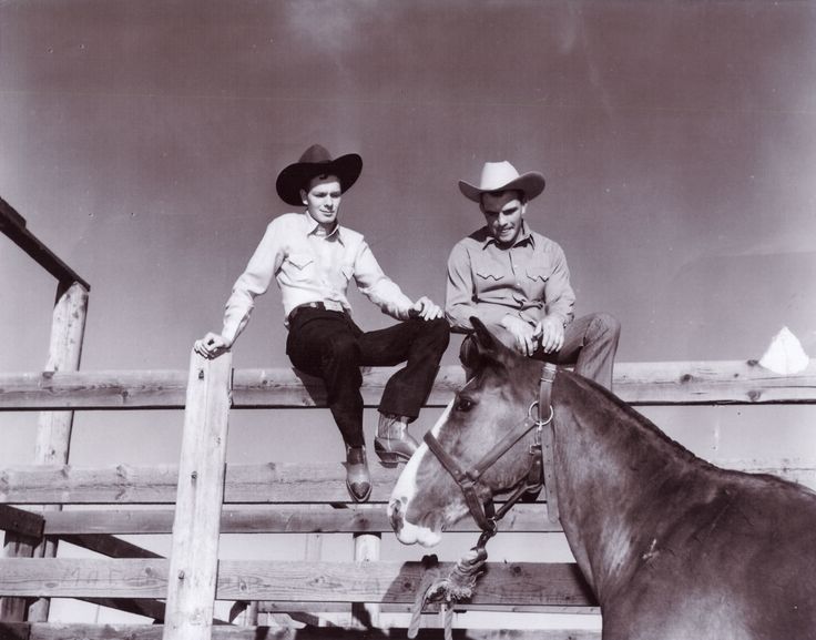 two men sitting on top of a wooden fence next to a horse