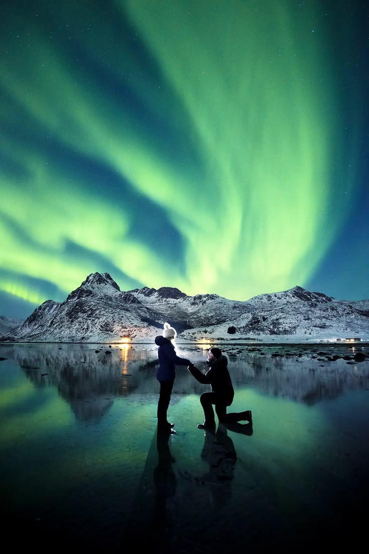 two people standing in front of a lake under the aurora lights